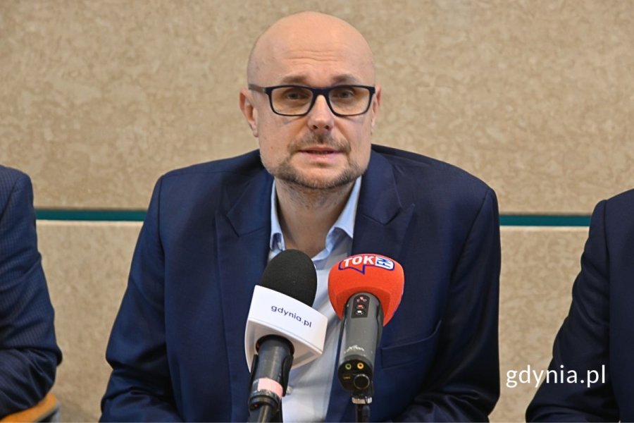 Mężczyzna w marynarce siedzący i mówiący do mikrofonu na tle beżowej ściany