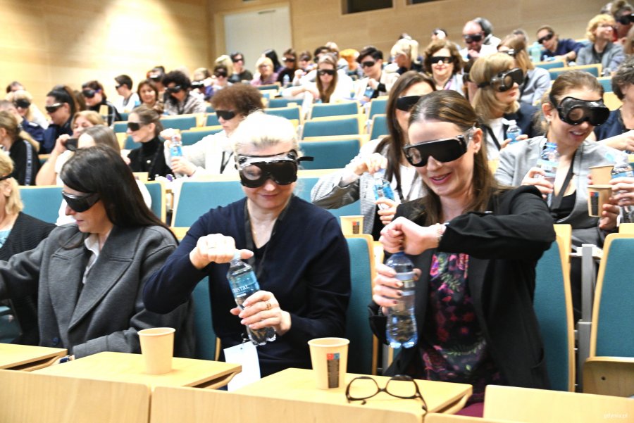 Osoby na widowni w czarnych okularach próbują otworzyć butelki w wodą