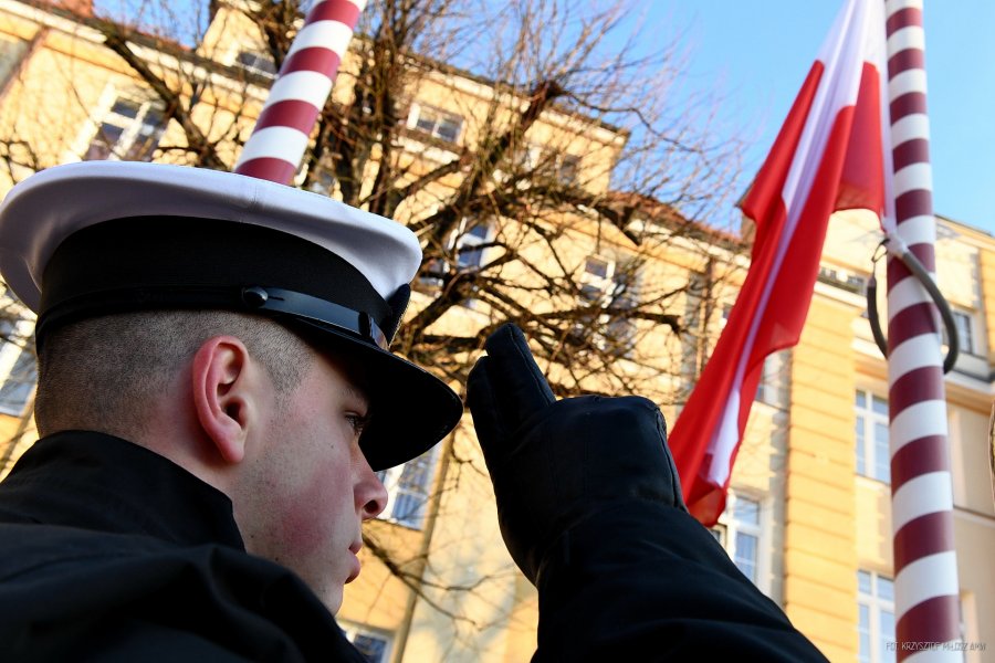 Głowa salutującego mężczyzny w marynarskiej czapce widziana od dołu. W tle wciągana na maszt flaga Polski.