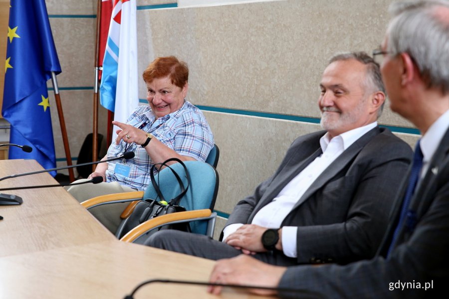 Trzy osoby siedzą za pulpitem w sali rady miasta.