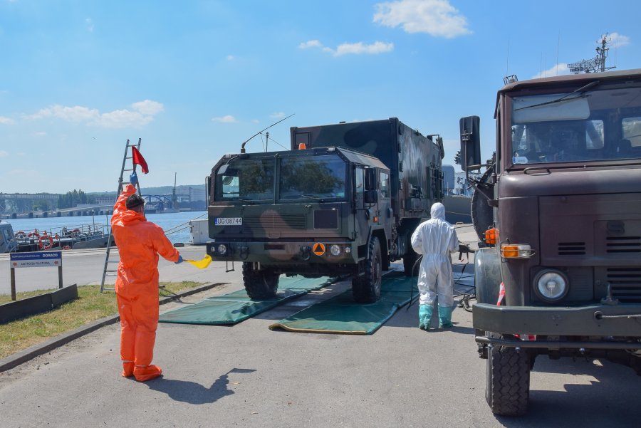 Sprzęt Morskiej Jednostki Rakietowej przy nabrzeżu portu Wojennego w Gdyni. Na pierwszym planie mężczyzna w pomarańczowym kombinezonie nawigujący ruchem. Dwie ciężarówki stoją przy nabrzeżu. Osoba w białym kombinezonie czyści i dezynfekuje jedną z nich // fot. por. mar. Cezary Skwierawski