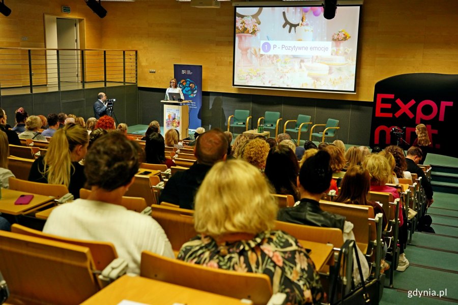 Na pierwszym planie uczestnicy konferencji. Za mównicą: dr Malwina Puchalska-Kamińska – psycholożka z Uniwersytetu SWPS wygłaszająca wystąpienie inauguracyjne podczas otwarcia konferencji „Dobrostan w centrum uwagi” (fot. Magdalena Czernek) 
