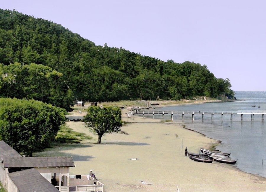 Plaża z łodziami rybackimi, z prawej strony morze i molo, z lewej zalesione wzgórza.