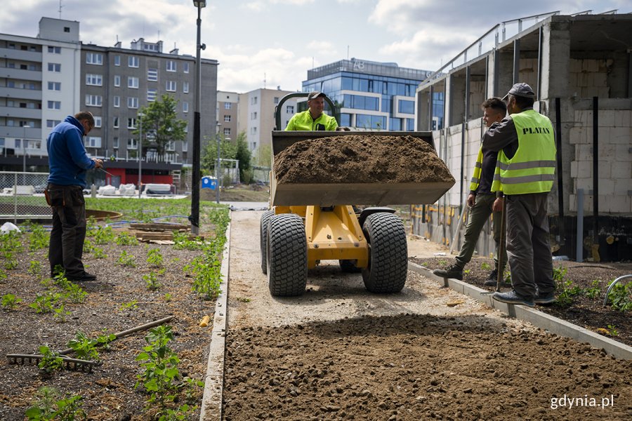 Koparka wiezie piasek, obok stoją robotnicy.