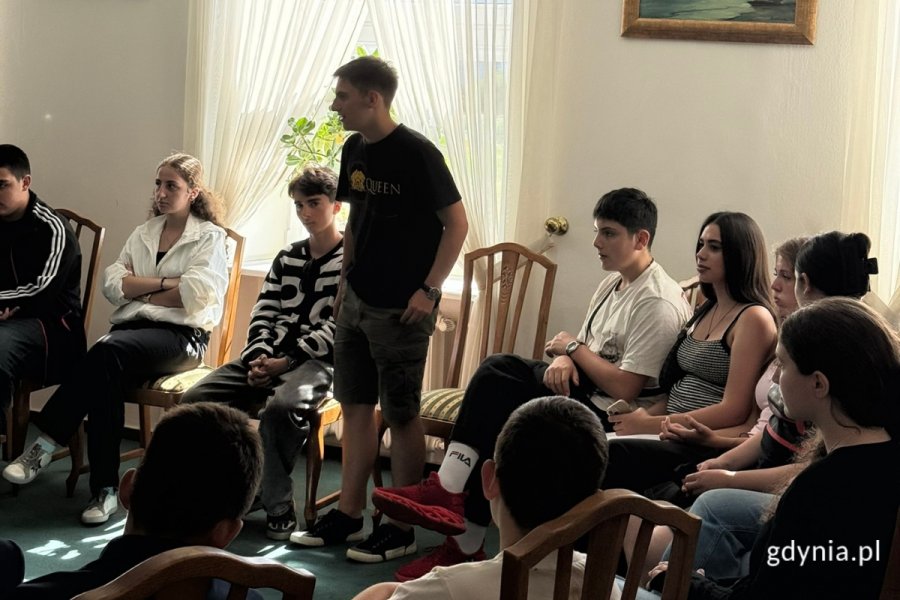 Przedstawiciel młodzieży z Gruzji w krótkich spodniach i czarnej koszulce przemawia podczas spotkania sali wizytowej, na fotelach wokół niego siedzi młodzież.