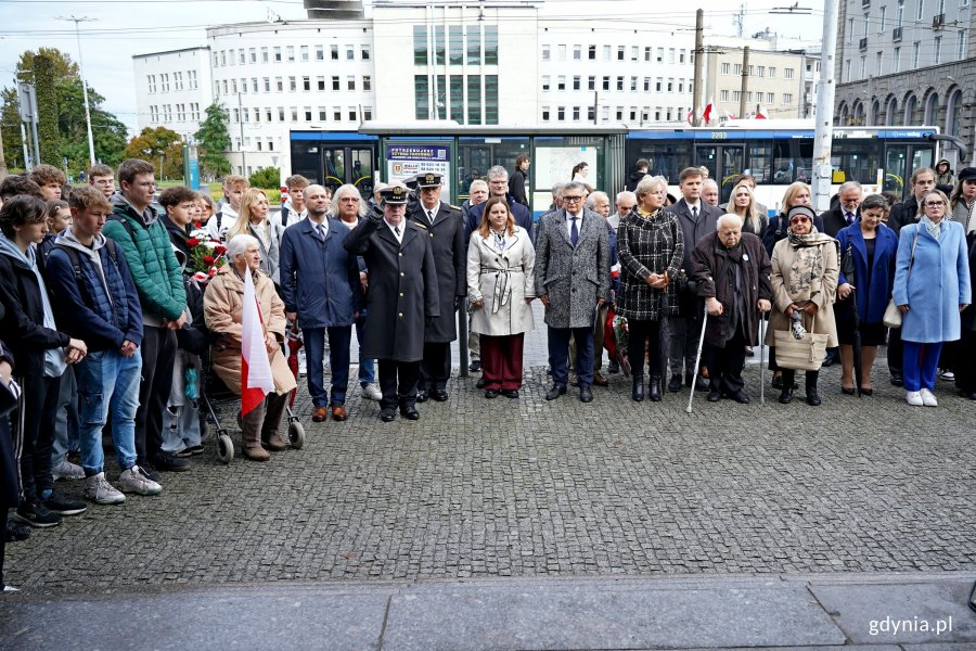 Uczestnicy uroczystości upamiętniających 86. rocznicę pierwszych wysiedleń gdynian zebrani przed tablicą pamiątkową na fasadzie budynku Dworca PKP Gdynia Główna