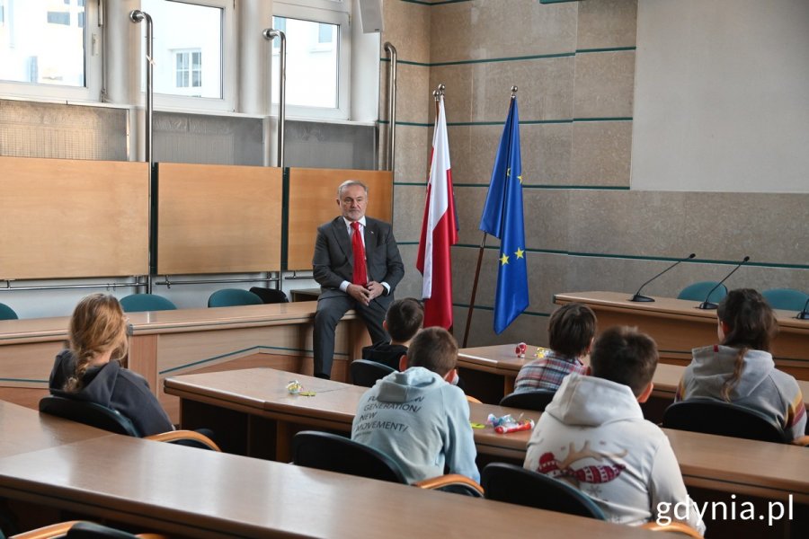 Wojciech Szczurek, prezydent Gdyni, na tle polskiej i unijnej flagi w sali obrad Rady Miasta Gdyni. Przed nim, za stołami siedzą uczniowie Szkoły Podstawowej nr 8. Zdjęcie ze spotkania w Urzędzie Miasta, które odbyło się 15 grudnia w ramach Światowego Tygodnia Przedsiębiorczości // fot. Magdalena Śliżewska