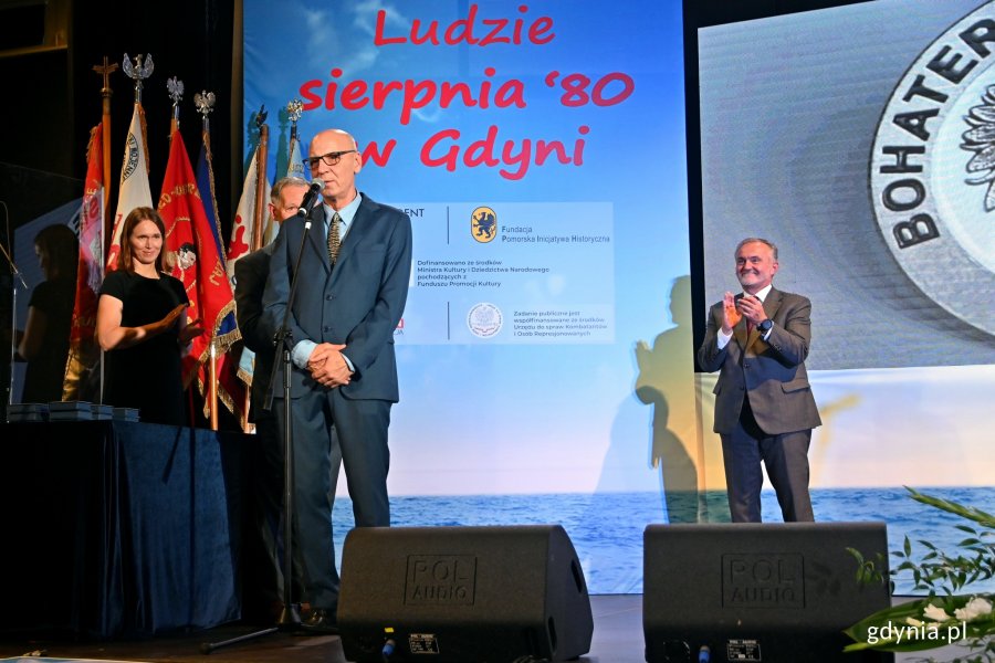 Andrzej Kołodziej, prezes Fundacji Pomorska Inicjatywa Historyczna, przemawia podczas uroczystości w sali koncertowej Zarządu Morskiego Portu Gdynia. Z tyłu - prezydent Gdyni Wojciech Szczurek bije brawo // fot. Magdalena Czernek
