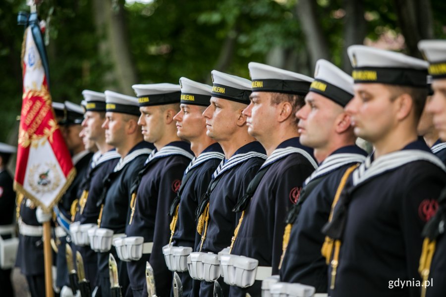 Żołnierze Marynarki Wojennej w mundurach stoją w rzędzie podczas uroczystości z okazji Dnia Sybiraka w Gdyni. Jeden z nich trzyma biało-czerwony sztandar