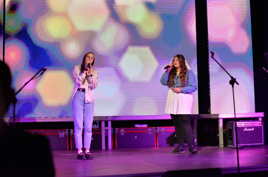 Dwie młode kobiety na scenie w trakcie wykonywania utworu muzycznego, w tle wyświetlany kolorowy obraz