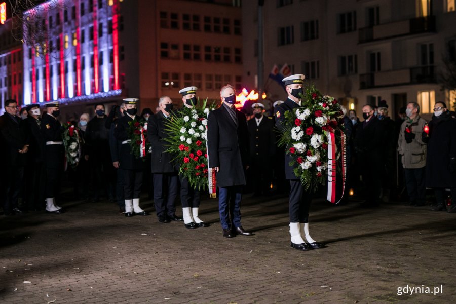 Uczestnicy uroczystości z okazji 50. rocznicy wydarzeń Grudnia '70 na placu Wolnej Polski składają wieńce. Na pierwszym planie marynarz z wieńcem składającym się z biało-czerwonych kwiatów i zielonych ozdobnych liści z szarfą z napisem Senat. Za nim stoi senator Sławomir Rybicki i kolejne delegacje // fot. Karol Stańczak