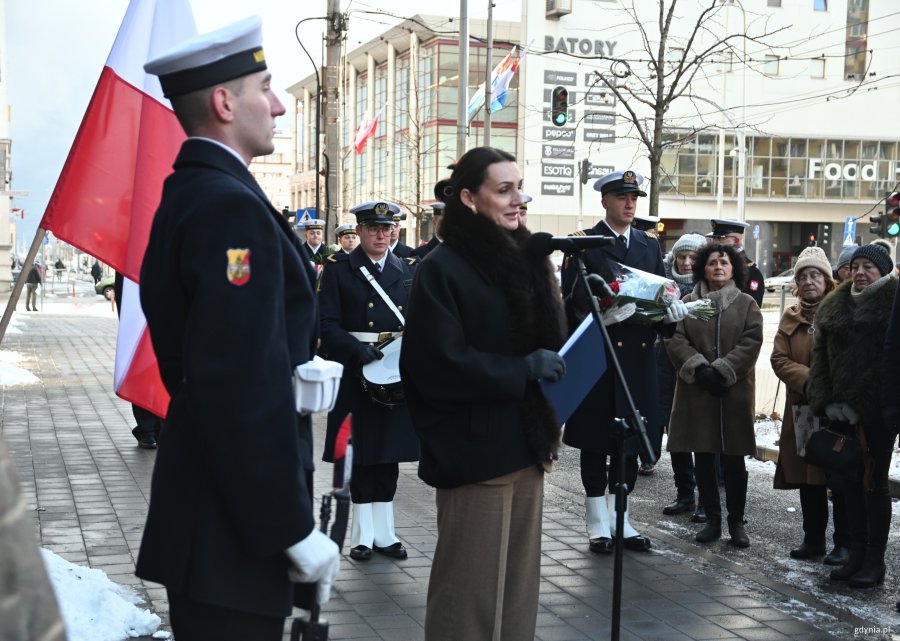 Kobieta w płaszczu przy mikrofonie, obok marynarz