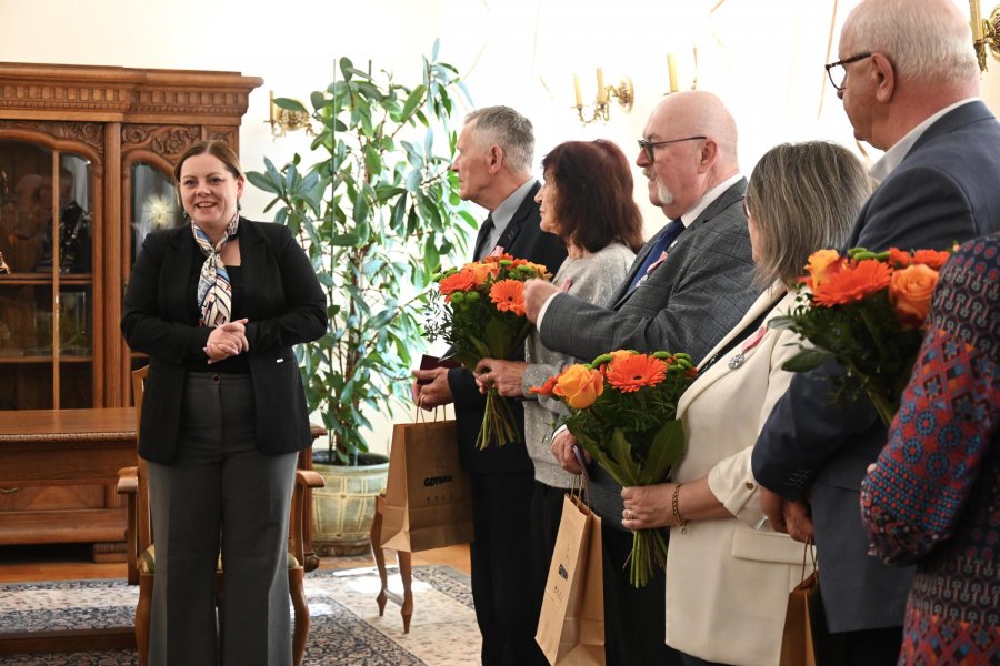 Kobieta w granatowej marynarce i grupa ludzi, kobiety mają pomarańczowe bukiety kwiat&oacute;w