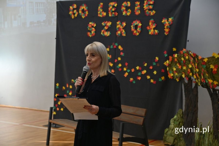 Stojąca kobieta mówiąca do mikrofonu w szkolnej hali sportowej