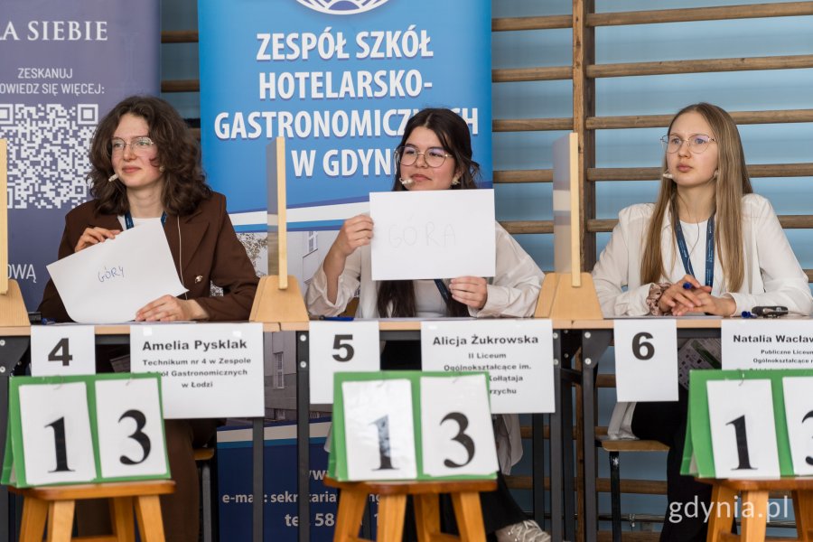 trzy kobiety siedzace przy stanowiskach konkursowych, trzymające kartki z odpowiedziami 