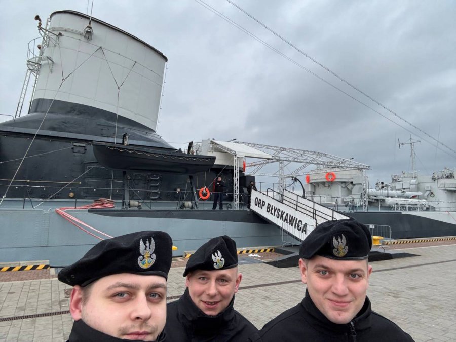Mężczyźni stojący na nabrzeżu na tle ORP „Błyskawica”, widoczny trap z nazwą okrętu