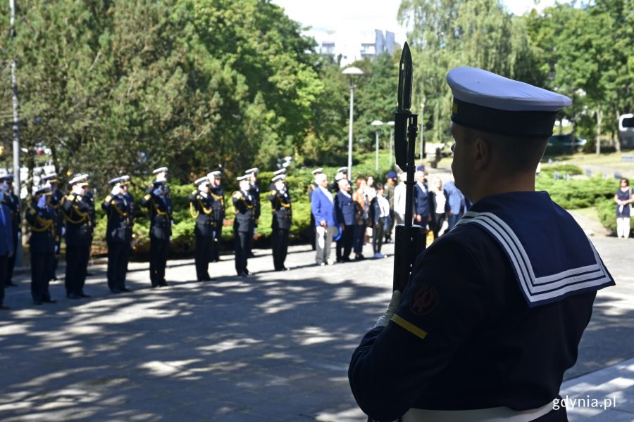 Marynarz stojący z bronią w ręku pełni wartę honorową pod pomnikiem podczas uroczystości na Cmentarzu Wojennym w Redłowie. W tle salutujący przedtawiciele Marynarki Wojennej