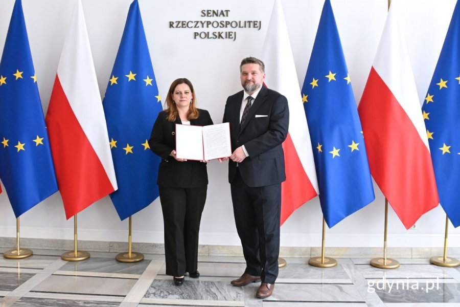 Prezydent Gdyni Aleksandra Kosiorek i przewodniczący Rady Miasta Gdyni Tadeusz Szemiot z pamiątkowym egzemplarzem uchwały stojący na tle flag Polski i Unii Europejskiej