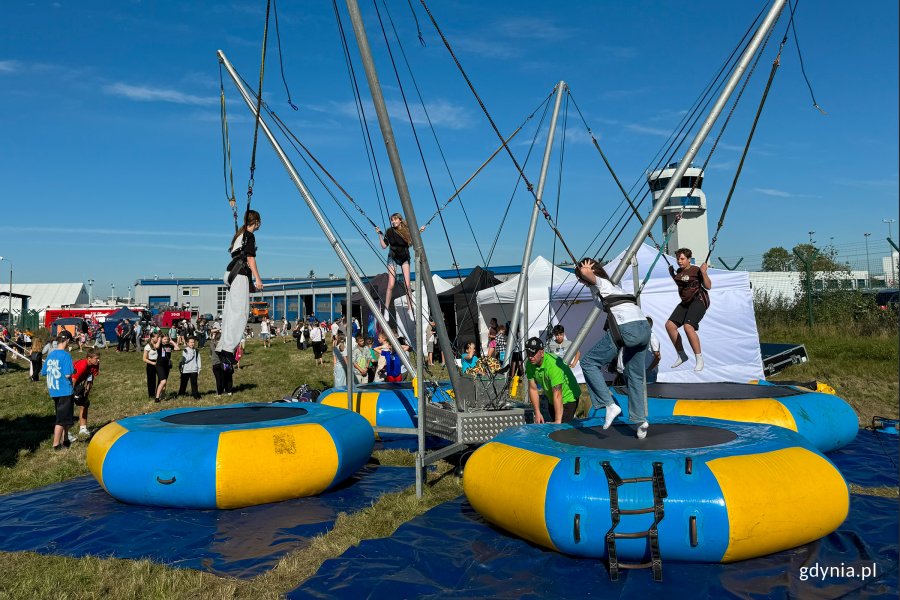 Młodzież skacząca na trampolinach w kolorze żółto-niebieskim w tle wieża portu lotniczego