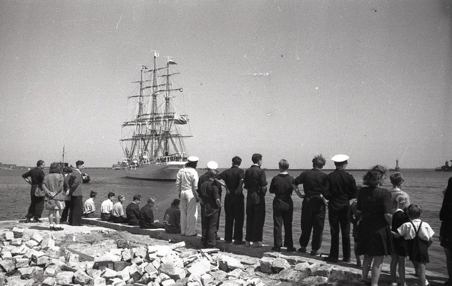 Negatyw czarno-biały z 1946 roku przedstawia tłum ludzi na zniszczonym nabrzeżu, którzy żegnają wypływający w rejs statek „Dar Pomorza”.  W głębi widoczna rufa płynącego żaglowca. Fotografia pochodzi z archiwum cyfrowego Muzeum Miasta Gdyni // fot. Edmund Zdanowski