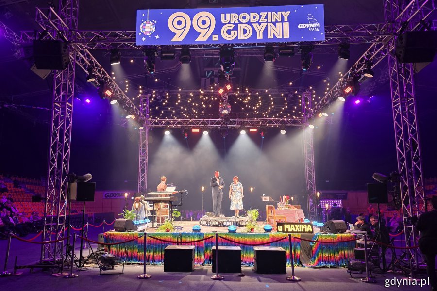 Hala Polsat Plus Arena Gdynia, scena, kolorowe oświetlenie, na scenie stoją wicewojewoda i prezydent Gdyni, napis "99. urodziny Gdyni" na wyświetlaczu