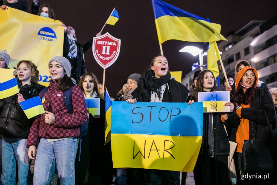 Mieszkańcy maszerują wieczorem ulicą podczas marszu antywojennego, trzymając flagi Ukrainy i hasła antywojenne.