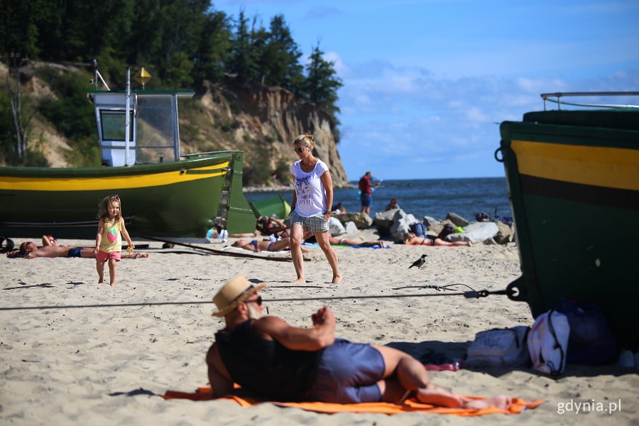 Plaża w Orłowie. Na pierwszym planie mężczyzna leży obok kutra rybackiego, dalej spaceruje kobieta z dzieckiem, na kolejnym planie następny kuter rybacki i plażowicze, w tle porośnięty drzewami Klif Orłowski i morze. Fot, Przemysław Kozłowski