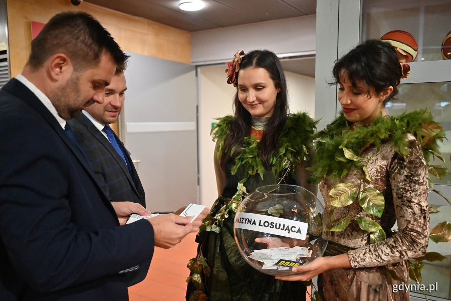 Kobiety trzymają szklaną kulę z losami i napisem: maszyna losująca, a panowie wyciągają losy