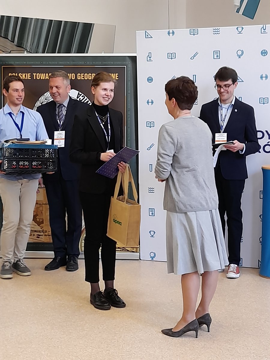 Nagrodę dla najlepszej laureatki wręczyła Ewa Śliwińska, stojąca tyłem do obiektywu, dyrektor gdyńskiego VI LO. W tle dwóch uczniów z nagrodami, ścianka III LO i Polskiego Towarzystwa Geograficznego.