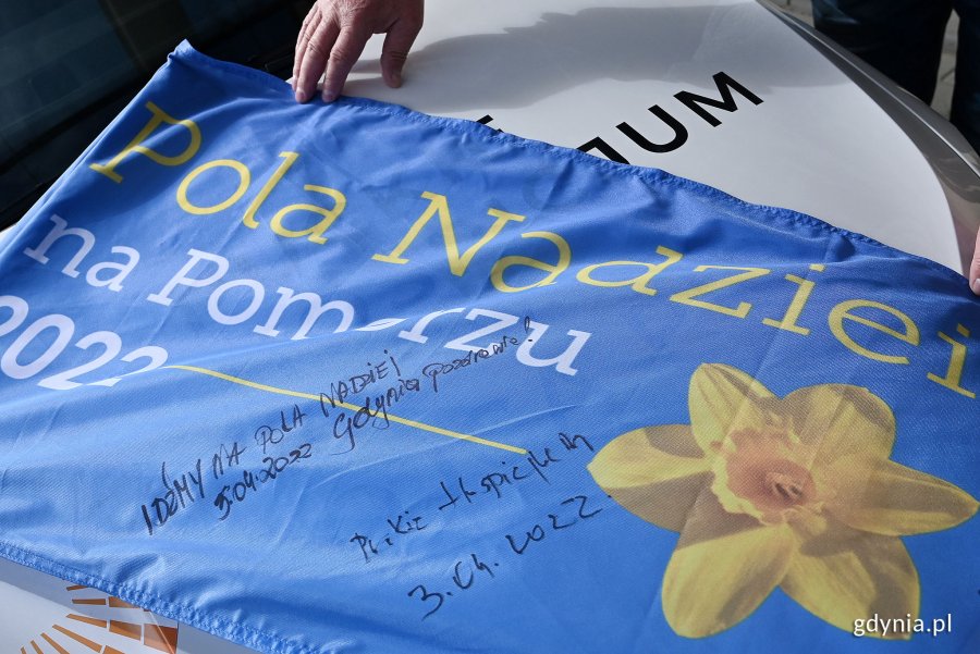 Niebieska flaga ze zdjęciem żonkila i napisem: „Pola Nadziei na Pomorzu 2022”. Są na niej dwa podpisy dopisane czarnym markerem: „Idźmy na Pola Nadziei . 3.04.2022, Gdynia pozdrawia” oraz „Puckie hospicjum - 3.04.2022” // fot. Michał Puszczewicz
