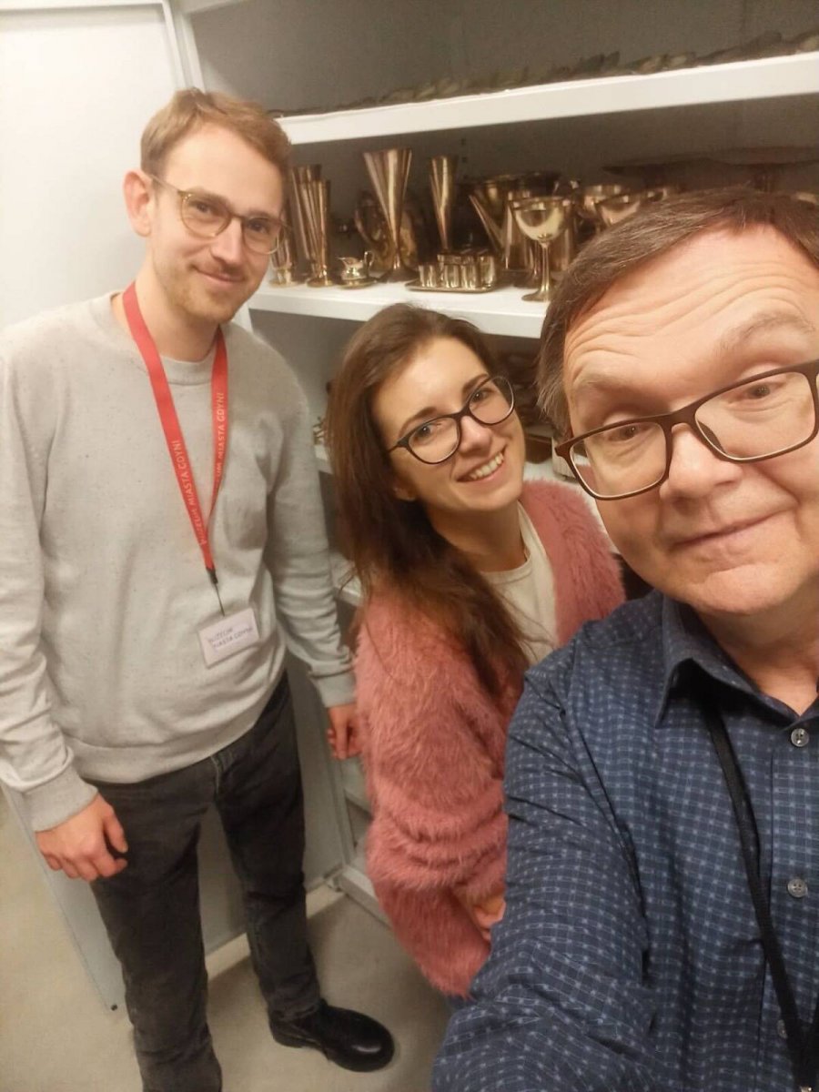 Mężczyźni i kobieta na tle regału wypełnionego starymi kielichami i pucharami