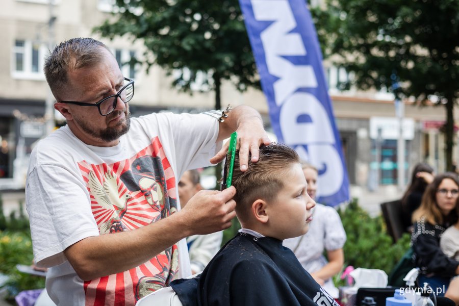 Mężczyzna strzyże chłopca siedzącego na fotelu fryzjerskim ustawionym na chodniku.