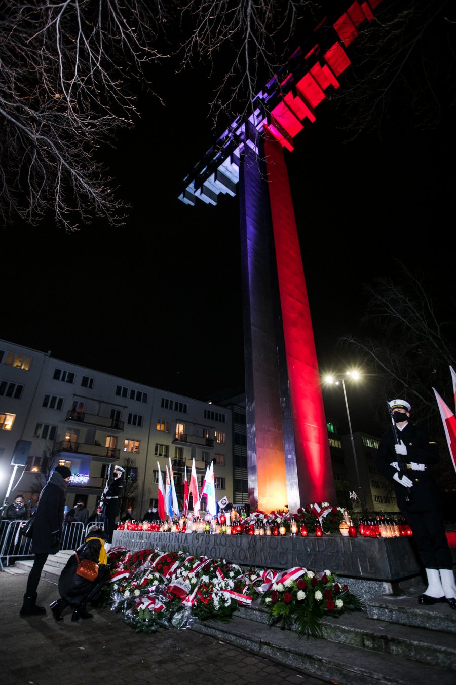Pomnik Ofiar Grudnia '70 na placu Wolnej Polski . Krzyż oświetlony w barwach narodowych. Przed pomnikiem dwie kobiety - jedna składa kwiaty, druga stoi. Po obu stronach pomnika posterunek honorowy złożony z marynarzy. Przed pomnikiem leży mnóstwo wieńców i wiązanek kwiatów. Płyta pomnika wypełniona jest zniczami // fot. Karol Stańczak