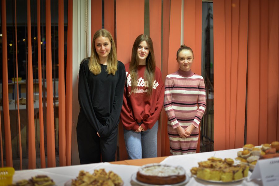 Trzy dziewczęta stojące na tle okna, na pierwszym plane stół z wypiekami