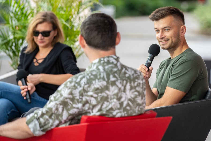 Po lewej Barbara Klicka w ciemnej marynarce oraz czarnych okularach, na środku Jakub Małecki - autor książki "Saturnin" - w zielonym t-shircie oraz po prawej Maciej Jakubowiak we wzorzystej koszuli siedzą w Muszli na placu Grunwaldzkim w Gdyni podczas spotkania w ramach festiwalu "Miasto Słowa" // fot. Anna Rezulak