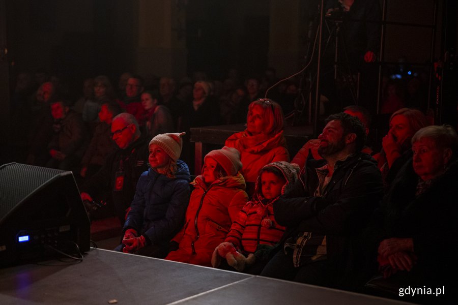 Publicznośc siedzi w ławkach w kościele, wśród nich trójka dzieci.
