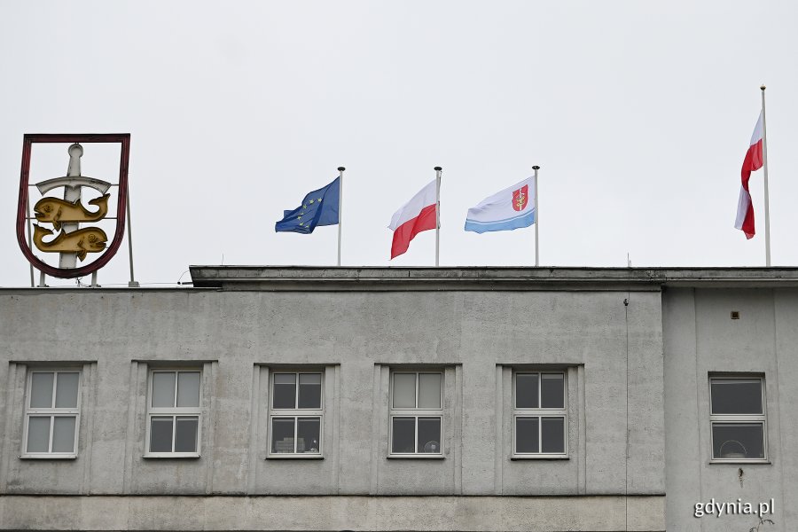 Flagi powiewające na budynku Urzędu Miasta Gdyni (od lewej): flaga Unii Europejskiej, flaga Polski, flaga Gdyni. Na maszcie biało-czerwona flaga. Po lewej stronie podświetlany herb Gdyni // fot. Michał Puszczewicz