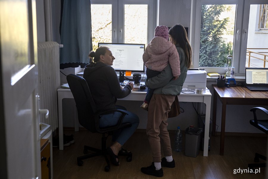 Kobieta siedzi przy biurku za komputerem, obok stoi kobieta z dzieckiem na ręku.