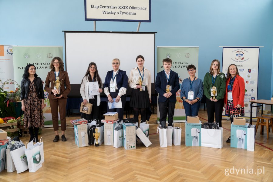dziewięć osób stojących na sali gimnastycznej, trzymają w rękach puchary, obok nich nagrody rzeczowe w torbach 