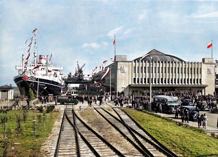 Na pierwszym planie tory kolejowe, które wiodą do budynku dworca morskiego, obok którego cumuje statek.