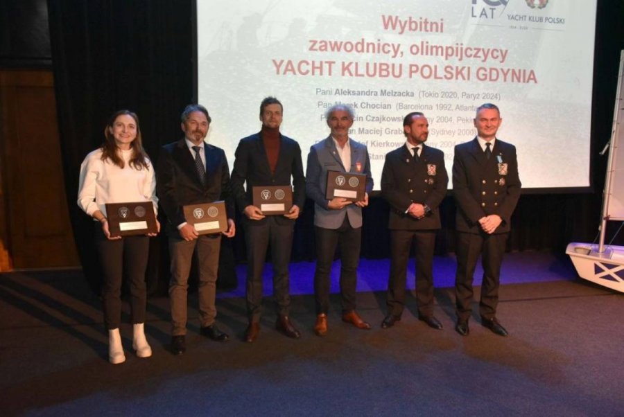Grupa osób stojących na scenie i mających za plecami biały ekran