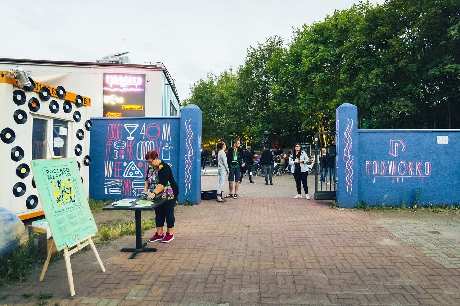 Wejście na patio Podwórka Art przy Klubie Muzycznym "Ucho", gdzie odbył się pokaz jednego z przedstawień inaugurujących festiwal „Pociąg do Miasta”. Na środku - grupa osób stojących przed wejściem. Po lewej - zielony baner promujący festiwal // fot. Krzysztof Winciorek