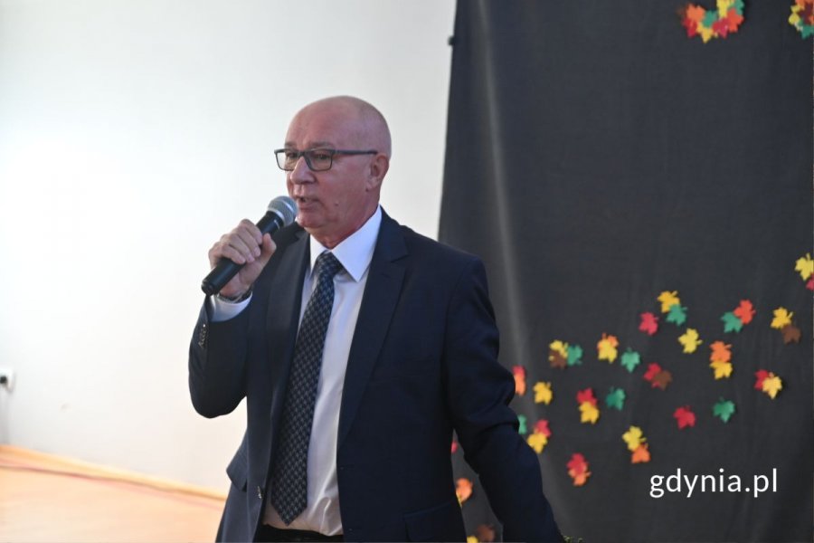 Stojący mężczyzna mówiący do mikrofonu w szkolnej hali sportowej