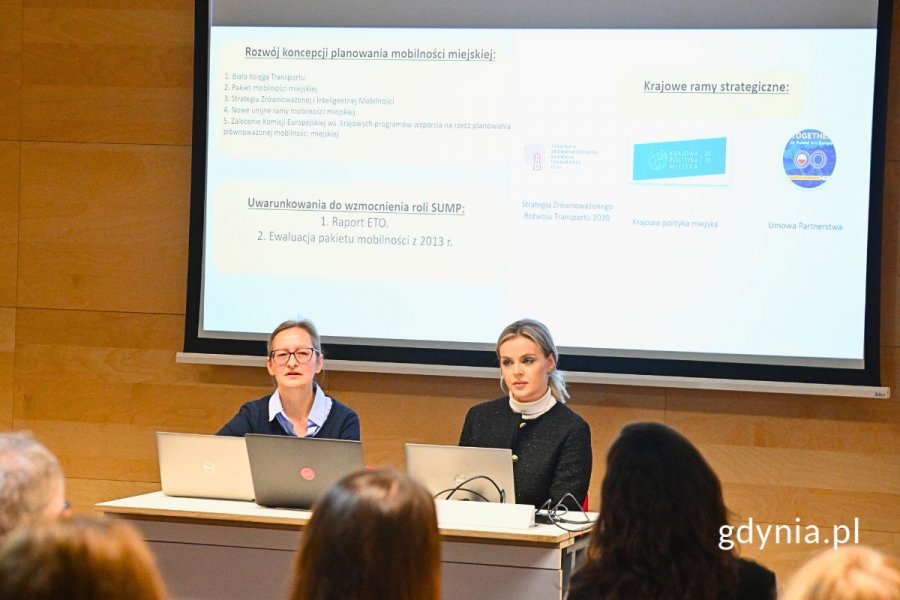Dwie kobiety siedzące przy stole, na którym stoją laptopy, za nimi widoczny ekran z wyświetloną prezentacją