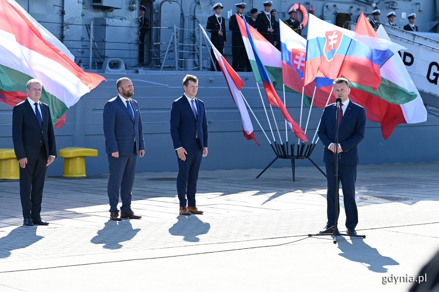 Od lewej: Tibor Benkő - minister obrony Węgier, Jaroslav Naď - minister obrony Słowacji oraz Jan Havránek - wiceminister obrony Czech. Przy mikrofonie przemawia Mariusz Błaszczak - minister obrony narodowej. W tle powiewają flagi Polski, Węgier, Słowacji i Czech. Za nimi okręt ORP "Gen. K. Pułaski", na którym ustawieni z szeregu stoją marynarze // fot. Michał Puszczewicz