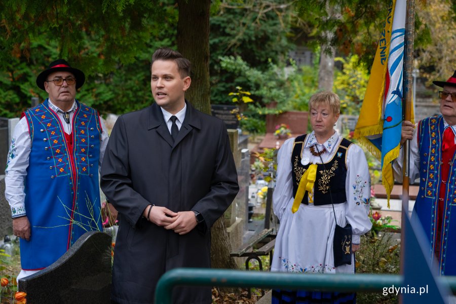 Mężczyzna w czarnym garniturze i para w regionalnym stroju nad grobem.