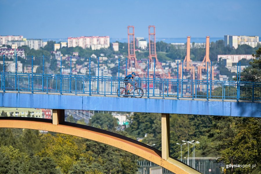 Kolarz-mężczyzna podczas przejazdu przez Gdynię, w tle widoczne zabudowania i port Gdynia. 