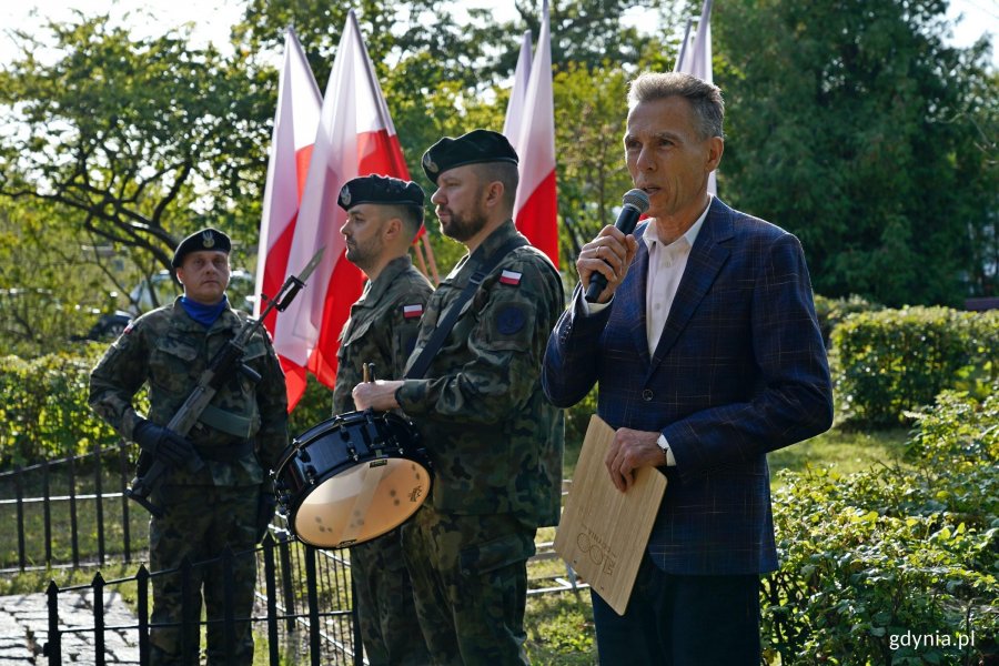 Krzysztof Dąbrowski prowadzący uroczystości z okazji 87. rocznicy zakończenia walk w obronie Kępy Oksywskiej. Obok niego trębacz i werblista oraz żołnierz stojący na posterunku honorowym przy Obelisku Obrońc&oacute;w Kępy Oksywskiej na Babich Dołach 