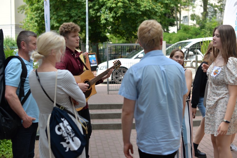 Grupa osób pod szkołą , w tle żagle z logo festiwalu i schody do szkoły, chłopak gra na gitarze