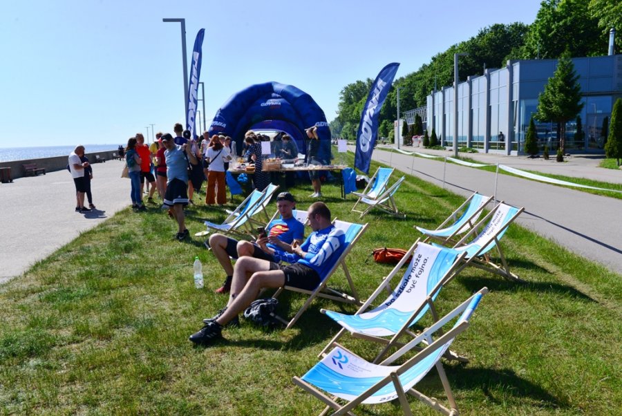 Bulwar, na środku trawa, na której stoją lerzaki w biało-błękitnych barwach, w tle niebieski dmuchany namiot Gdynia Sport. Na leżakach odpoczywa dwóch biegaczy, w tle stoisko ze słodkim kiermaszem III LO, przy nim grupka ludzi